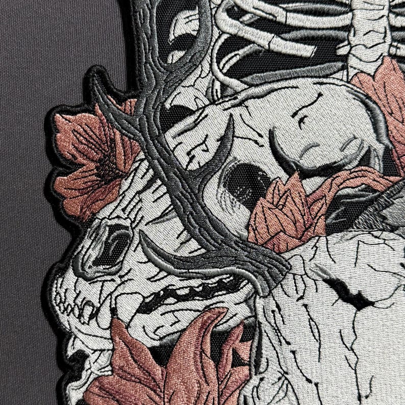 May include: Embroidered patch featuring a detailed skull with antlers and floral accents in shades of white, gray, and coral. The patch has a black border and is likely for jackets or bags.