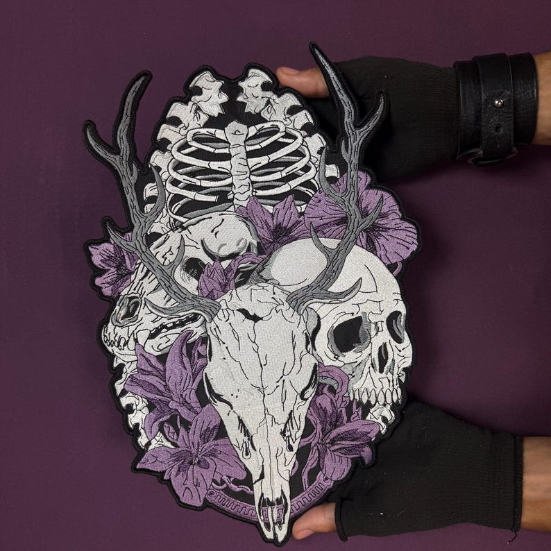 Op de afbeelding: Een grote geborduurde patch met een centrale hertenkop met gewei, geflankeerd door een menselijke schedel en een ribbenkast. Het ontwerp is geaccentueerd met paarse bloemen en bladeren, uitgevoerd in wit, grijs en paars garen op een zwarte achtergrond.