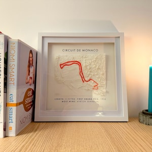 May include: A framed 3D map of the Circuit de Monaco race track, with a red track outline. The frame is white, and the text "CIRCUIT DE MONACO" is visible. A teal candle burns beside the frame, and several books are on the left.
