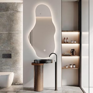 Op de afbeelding: Modern badkamerontwerp met een grote, organisch gevormde spiegel met een gloeiende rand. Een zwarte marmeren wastafel staat op een houten voetstuk, aangevuld met een zwarte kraan. Plankjes met toiletartikelen en een toilet maken de minimalistische esthetiek compleet.