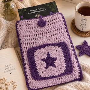 Pode incluir: Uma capa de livro em crochê lilás com um remendo quadrado roxo escuro com um motivo de estrela central. A capa inclui um fecho de botão roxo escuro no topo. Um livro está parcialmente inserido na capa, mostrando o texto Haunted. Hunter. Destined for each other.