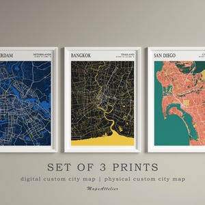 Peut inclure: Ensemble de trois impressions encadrées de cartes de villes. Les cartes présentent Amsterdam en bleu, Bangkok en noir et jaune, et San Diego en rouge et vert. Le texte "SET OF 3 PRINTS" est en dessous.