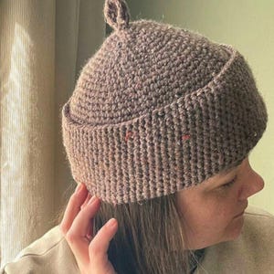 May include: A brown, textured, hand-crocheted beanie hat with a small, rounded knob at the very top. The hat features a thick, folded brim and is crafted from a yarn that includes subtle, scattered flecks of orange and dark brown throughout.
