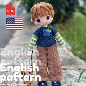 以下が含まれることがあります： 手作りの茶色の髪の毛の編み人形。青いセーターに花のディテール、茶色のズボン、ストライプの袖を着ています。人形は、小さな編み犬に付けられたリードを持っています。画像には「English pattern」というテキストが含まれています。