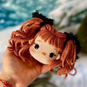 Op de afbeelding: Een gehaakte poppenkop met bruin haar, zwarte haarbandjes en een lichtbruin gezicht. De pop heeft grote zwarte ogen en een kleine roze neus.