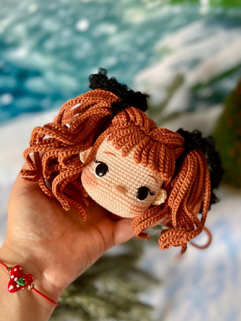 Op de afbeelding: Een gehaakte poppenkop met bruin haar, zwarte haarbandjes en een lichtbruin gezicht. De pop heeft grote zwarte ogen en een kleine roze neus.
