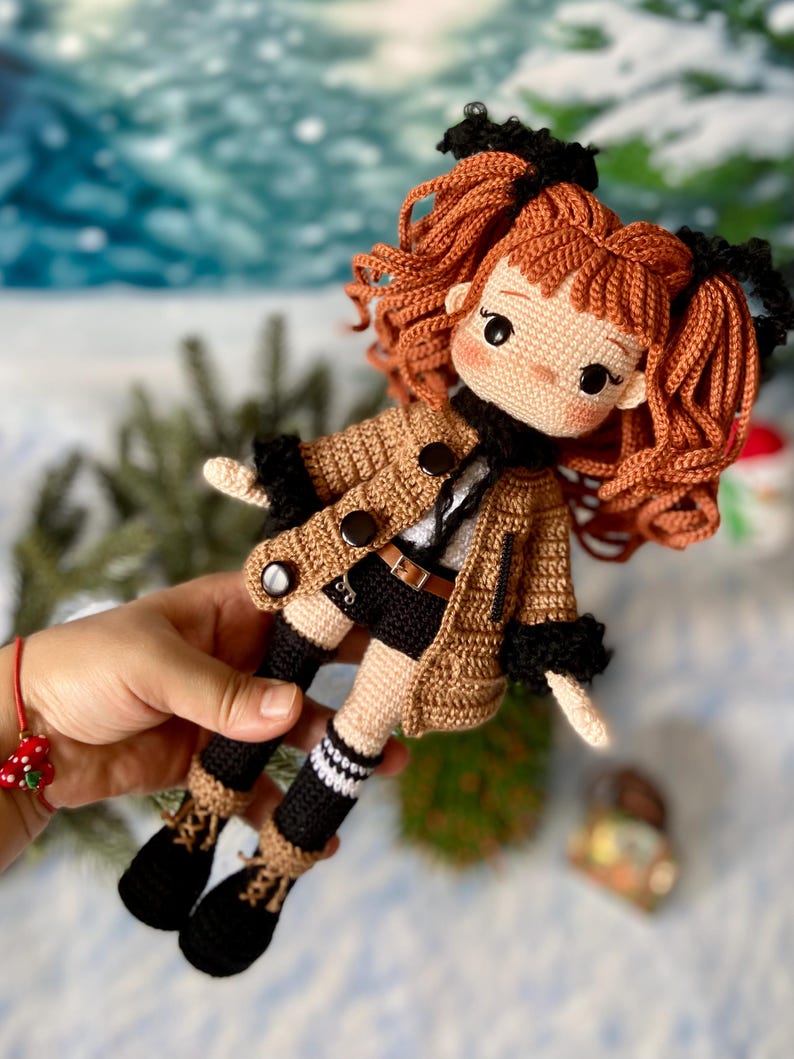 Op de afbeelding: Een gehaakte pop met bruin haar, die een bruin jasje, een zwarte broek en zwarte laarzen draagt. De pop heeft een zwarte riem en witte strepen op de sokken.