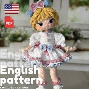 Könnte beinhalten: Eine handgefertigte Häkelpuppe mit blonden Haaren, die ein weißes Kleid mit rosa und blauen Akzenten trägt. Die Puppe hat einen süßen Ausdruck und ist mit einer Schleife und Perlen verziert. Die Worte "English pattern" sind sichtbar.