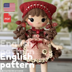 Puede incluir: Muñeca de ganchillo hecha a mano con sombrero y vestido rojos, con un delantal rosa adornado con una flor y un lazo. La muñeca tiene cabello castaño y pendientes decorativos. Incluye el texto "English pattern".