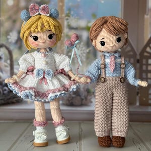 Puede incluir: Dos muñecos de ganchillo hechos a mano, una niña y un niño, están uno al lado del otro. La muñeca de la niña tiene el pelo rubio, un vestido blanco con detalles rosas y azules y zapatos blancos. El niño tiene el pelo castaño, una camisa azul y pantalones marrones. Se dan la mano.