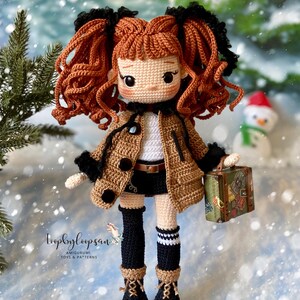 Op de afbeelding: Gehaakte pop met bruin krullend haar, die een bruine jas, zwarte leggings, witte sokken en zwarte laarzen draagt. De pop houdt een kleine koffer met reisstickers vast. De pop staat op een besneeuwde achtergrond.