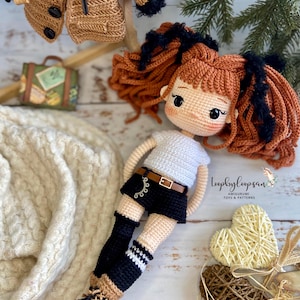 Op de afbeelding: Gehaakte amigurumi pop met bruin haar, zwarte ogen en een wit shirt. De pop draagt een zwarte broek, zwart-wit gestreepte sokken en zwarte schoenen. De pop zit op een wit houten oppervlak met twee geweven harten.