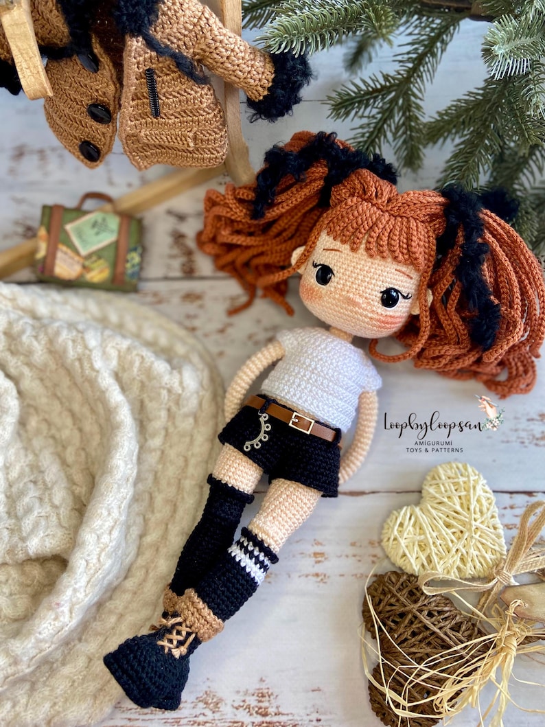 Op de afbeelding: Gehaakte amigurumi pop met bruin haar, zwarte ogen en een wit shirt. De pop draagt een zwarte broek, zwart-wit gestreepte sokken en zwarte schoenen. De pop zit op een wit houten oppervlak met twee geweven harten.