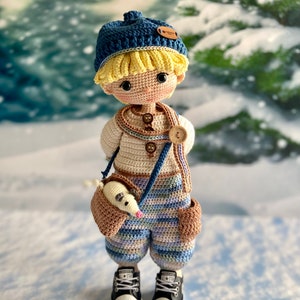 K&ouml;nnte beinhalten: Eine geh&auml;kelte Puppe mit einem blauen Hut, einem cremefarbenen Pullover, einer gestreiften Hose und schwarzen Turnschuhen. Die Puppe h&auml;lt ein kleines geh&auml;keltes Tier in einer braunen Tasche.