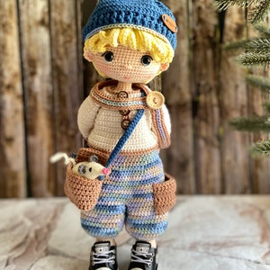 K&ouml;nnte beinhalten: Eine geh&auml;kelte Puppe mit einem blauen Hut, einem wei&szlig;en Pullover, einer gestreiften Hose und schwarzen Turnschuhen. Die Puppe h&auml;lt eine braune geh&auml;kelte Tasche mit einem kleinen wei&szlig;en geh&auml;kelten Tier darin.