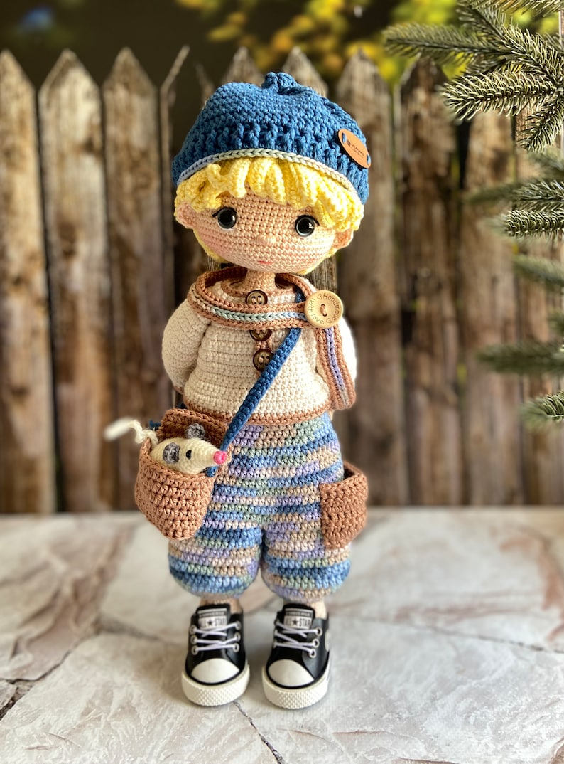 K&ouml;nnte beinhalten: Eine geh&auml;kelte Puppe mit einem blauen Hut, einem wei&szlig;en Pullover, einer gestreiften Hose und schwarzen Turnschuhen. Die Puppe h&auml;lt eine braune geh&auml;kelte Tasche mit einem kleinen wei&szlig;en geh&auml;kelten Tier darin.