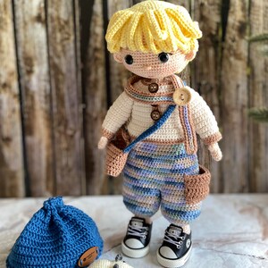 K&ouml;nnte beinhalten: Eine geh&auml;kelte Puppe mit blonden Haaren, die ein blau-braunes gestreiftes Outfit, braune Schuhe und eine braune Tasche tr&auml;gt. Die Puppe steht neben einem blauen H&auml;kelhut und einer wei&szlig;en H&auml;kelmaus.