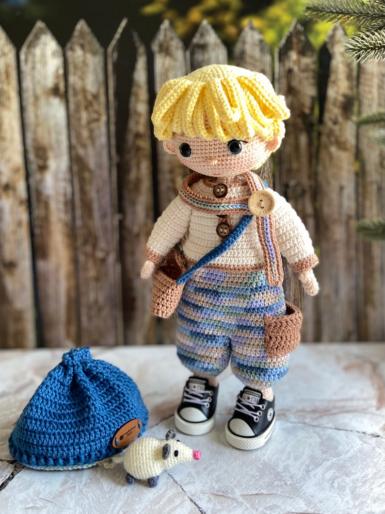 K&ouml;nnte beinhalten: Eine geh&auml;kelte Puppe mit blonden Haaren, die ein blau-braunes gestreiftes Outfit, braune Schuhe und eine braune Tasche tr&auml;gt. Die Puppe steht neben einem blauen H&auml;kelhut und einer wei&szlig;en H&auml;kelmaus.