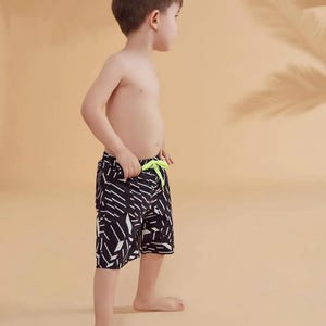 Peut inclure: Un jeune gar&ccedil;on portant un short de bain &agrave; motifs noirs et blancs avec un cordon de serrage vert fluo. Le short pr&eacute;sente un motif g&eacute;om&eacute;trique. L'arri&egrave;re-plan est de couleur beige uni avec une ombre de feuille de palmier.