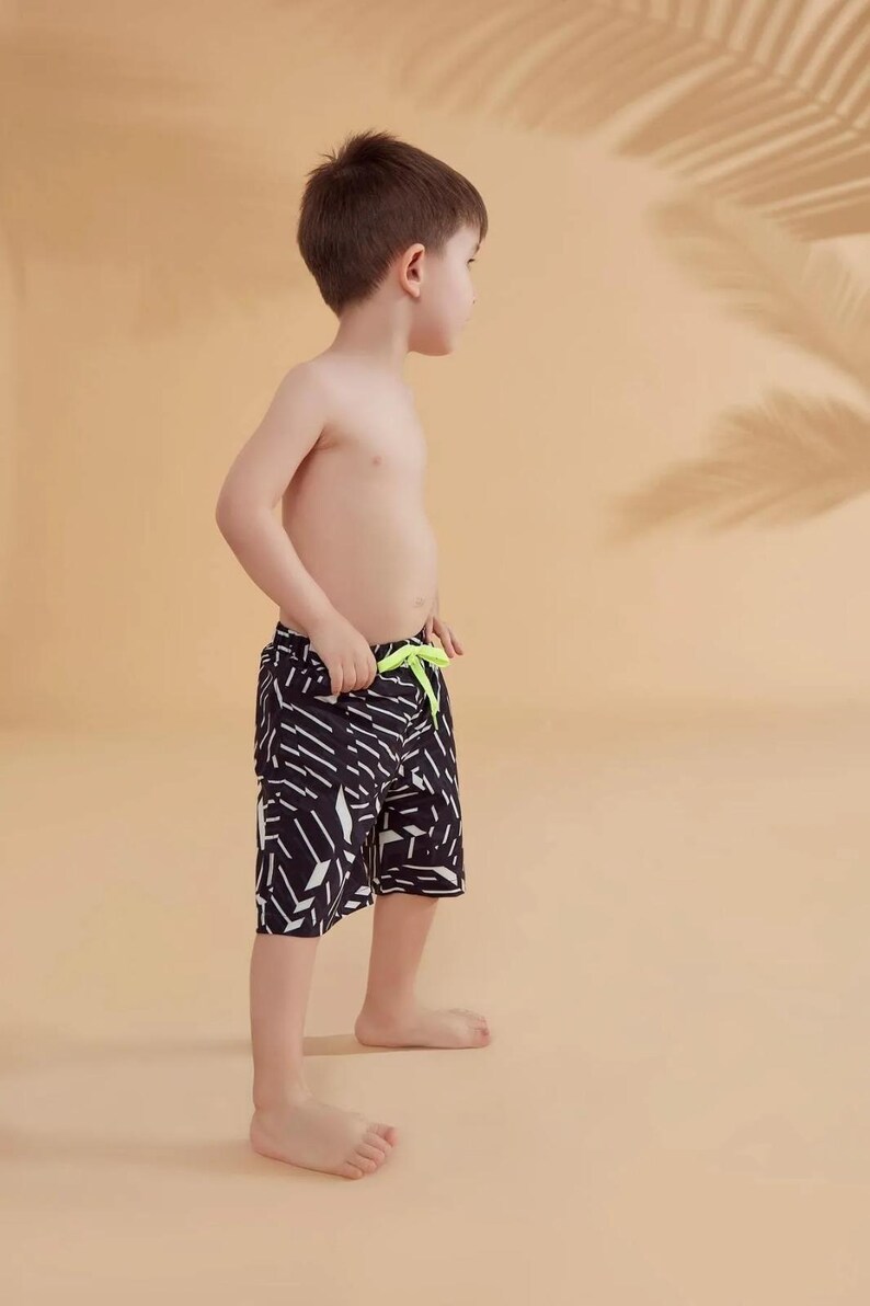 Peut inclure: Un jeune gar&ccedil;on portant un short de bain &agrave; motifs noirs et blancs avec un cordon de serrage vert fluo. Le short pr&eacute;sente un motif g&eacute;om&eacute;trique. L'arri&egrave;re-plan est de couleur beige uni avec une ombre de feuille de palmier.