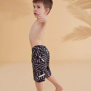 Peut inclure: Un enfant porte un short de bain &agrave; motifs noirs et blancs avec un cordon de serrage vert fluo. L'enfant est debout pieds nus, le bras lev&eacute;, sur un fond neutre.