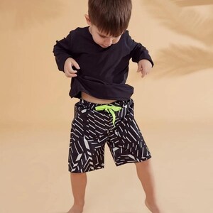 Peut inclure: Un enfant porte un haut noir &agrave; manches longues et un short &agrave; motifs noir et blanc avec un cordon de serrage vert fluo. Le short pr&eacute;sente un motif g&eacute;om&eacute;trique. L'enfant se tient sur un fond beige.