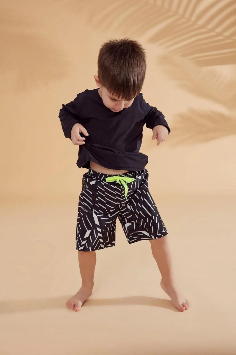Peut inclure: Un enfant porte un haut noir &agrave; manches longues et un short &agrave; motifs noir et blanc avec un cordon de serrage vert fluo. Le short pr&eacute;sente un motif g&eacute;om&eacute;trique. L'enfant se tient sur un fond beige.