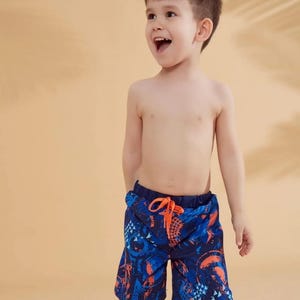 Peut inclure: Un jeune enfant portant un short de bain &agrave; motifs bleus et oranges. Le short a une taille avec cordon de serrage et un design vibrant. L'enfant sourit et regarde sur le c&ocirc;t&eacute;. L'arri&egrave;re-plan est de couleur beige uni.