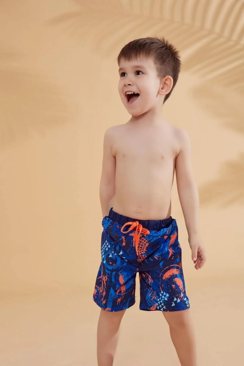 Peut inclure: Un jeune enfant portant un short de bain &agrave; motifs bleus et oranges. Le short a une taille avec cordon de serrage et un design vibrant. L'enfant sourit et regarde sur le c&ocirc;t&eacute;. L'arri&egrave;re-plan est de couleur beige uni.