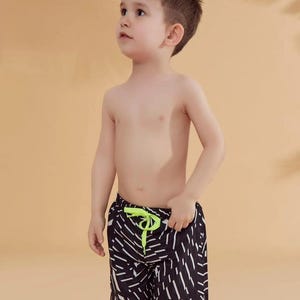Peut inclure: Un jeune enfant portant un short de bain &agrave; motifs noirs et blancs avec un cordon de serrage vert fluo. Le short a un motif g&eacute;om&eacute;trique. L'enfant regarde vers le haut.