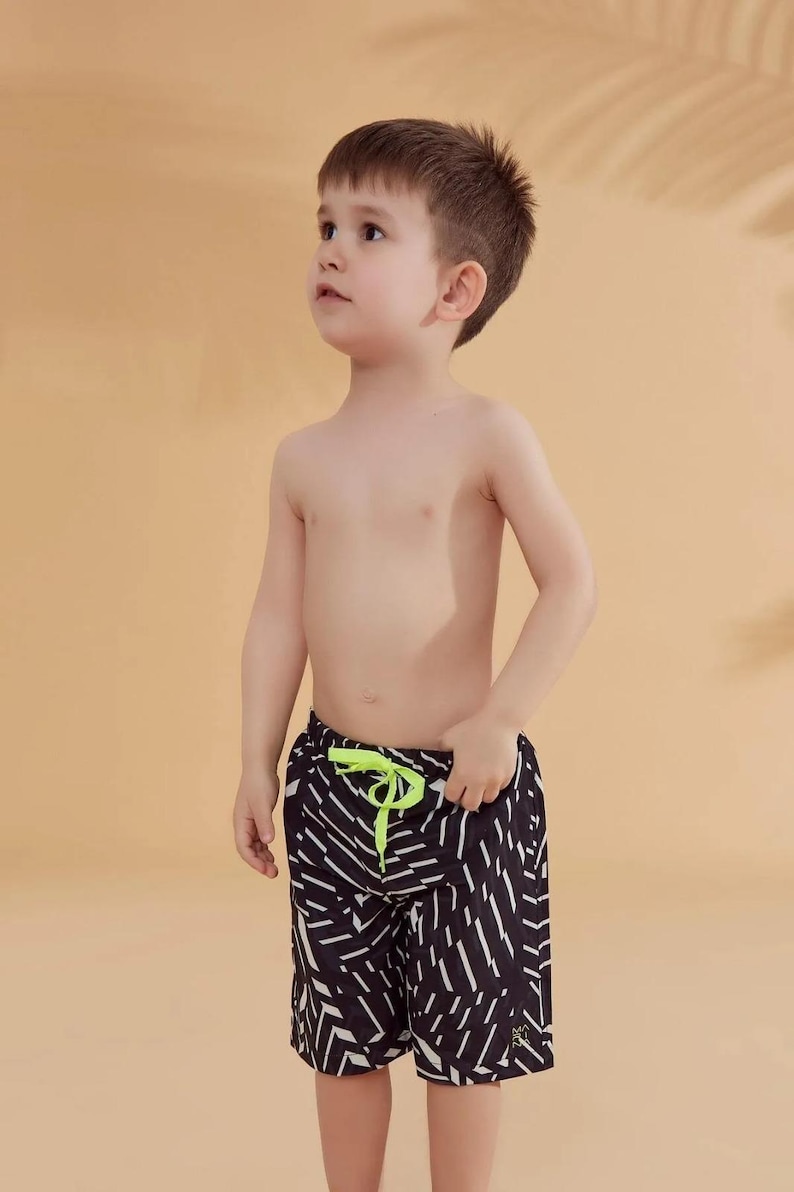 Peut inclure: Un jeune enfant portant un short de bain &agrave; motifs noirs et blancs avec un cordon de serrage vert fluo. Le short a un motif g&eacute;om&eacute;trique. L'enfant regarde vers le haut.
