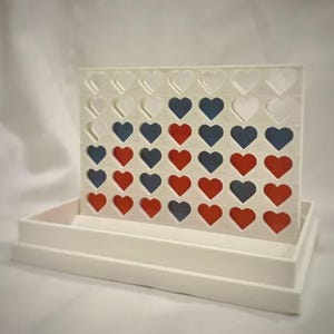 Op de afbeelding: Een wit Connect Four-spel met hartvormige openingen en rode, witte en blauwe speelstukken. Het bord staat op een witte lade. Dit tafelspel is geschikt voor alle leeftijden en is een leuke activiteit.