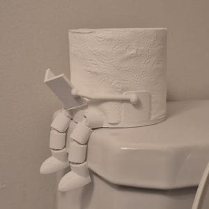 Op de afbeelding: Een witte toiletrolhouder met een grappig ontwerp. De houder heeft een rol wit toiletpapier met witte benen en een boek, waardoor het lijkt alsof een persoon leest terwijl hij op een toilet zit.