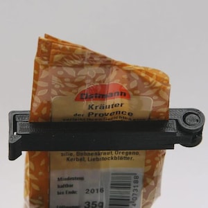 Op de afbeelding: Een zwarte plastic zakclip die een zak kruiden vasthoudt. De zak heeft een bloemenpatroon en de tekst "Kräuter der Provence". De zak bevat 35g kruiden.