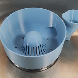 Op de afbeelding: Een lichtblauwe citruspersset, bestaande uit een kom met een centrale ruimer en een aparte tuit. De kom heeft een handgreepachtige uitsparing. De citruspers is gemaakt van plastic.