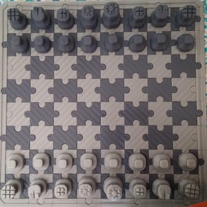 Op de afbeelding: Een grijs schaakspel met een uniek puzzelstukjesontwerp. Het bord bestaat uit in elkaar grijpende puzzelstukjes in licht- en donkergrijs. De schaakstukken zijn ook grijs, met verschillende vormen voor elk stuk. Een creatieve kijk op een klassiek spel.