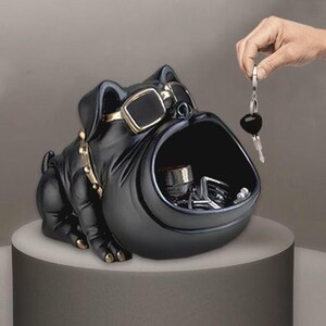Op de afbeelding: Een zwarte decoratieve opbergcontainer in de vorm van een bulldog met een grote open mond. De bulldog draagt een goudkleurige zonnebril en een gouden halsband. De container staat op een rond, lichtgekleurd oppervlak. Sleutels worden in de container geplaatst.