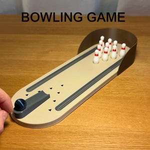 Op de afbeelding: Een miniatuur bowlingspel met de tekst "BOWLING GAME" bovenaan. Het spel heeft een beige baan met grijze bumpers en een donkerblauwe bal. Tien witte kegels met rode toppen staan aan het einde van de baan.