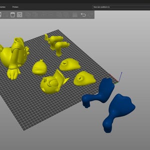 Puede incluir: Un modelo de pato de dibujos animados impreso en 3D, desmontado en varias piezas. Las piezas son amarillas y azules, y est&aacute;n dispuestas sobre una superficie de rejilla gris.