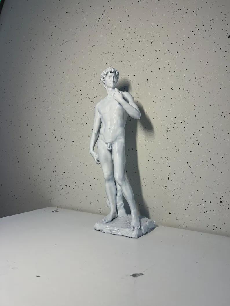 May include: A white, detailed sculpture of a nude male figure standing with one hand on his hip. The sculpture is on a white base.