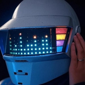 Peut inclure: Casque futuriste bleu clair avec une visière affichant une matrice de lumières bleues et des barres verticales colorées. Le casque a une finition lisse et mate sur fond sombre.