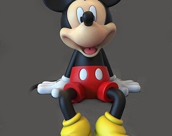 May include: A black and red Mickey Mouse figurine with yellow shoes. The figurine is sitting with its legs crossed and has a big smile on its face.
