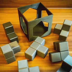 May include: A geometric puzzle game featuring a dark green cube frame and several modular blocks in shades of grey and green. The blocks are arranged around the frame on a wooden surface, suggesting a challenge or game.