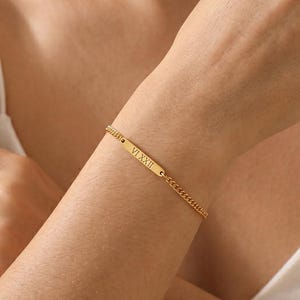 Könnte beinhalten: Ein goldfarbenes Kettenarmband mit einem zentralen rechteckigen Steg, der mit den römischen Ziffern VI XXII graviert ist. Das Armband besteht aus feinen ineinandergreifenden Gliedern und hat ein poliertes Metallfinish.
