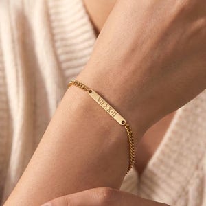 May include: Gold-colored bracelet with a rectangular bar and chain links. The bar is engraved with the Roman numeral VIIXXIII. The bracelet is worn on a wrist.