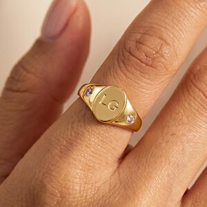 Può includere: Un anello sigillo dorato con una superficie ovale incisa con le lettere LG. L'anello è impreziosito da due piccole pietre rotonde di colore viola chiaro incastonate su entrambi i lati della superficie ovale centrale, creando un gioiello.