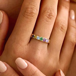 May include: A gold band ring featuring a row of six small, round, multi-colored gemstones including purple, green, light blue, yellow, and dark blue stones set horizontally across the center of the metal band.