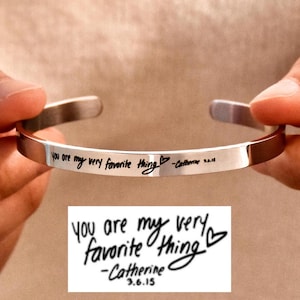 May include: A silver bangle bracelet with the inscription "You are my very favorite thing -Catherine 3.6.15" engraved on it. The inscription is a handwritten message.