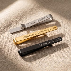 May include: Three metal tie clips in silver, gold, and black, each with engraved text. The silver clip reads "Forever Grateful X", the gold clip says "Groomsman - DAVID & SOPHIA - 2024", and the black clip says "The Groom - 10.12.24".