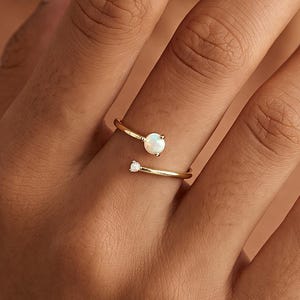 May include: A gold open-style ring featuring two round stones of different sizes. The larger stone is an iridescent opal, while the smaller stone is a clear, brilliant-cut gem. The band is thin, polished gold, creating a delicate and refined appearance.